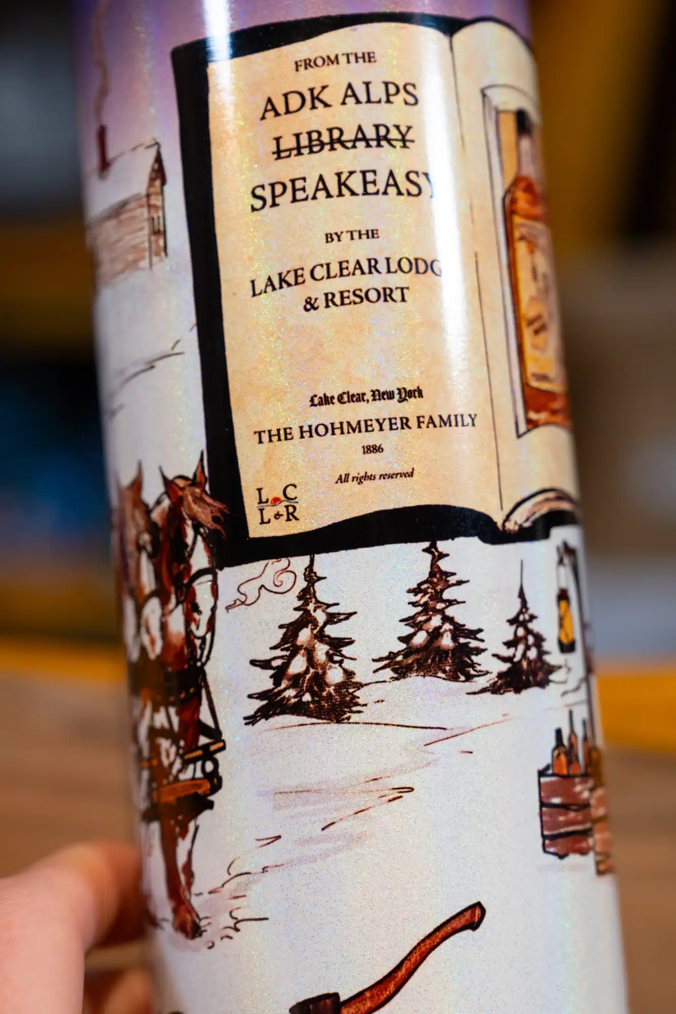 Lake Clear Lodge Holographic Tumbler detail with 'ADK Alps Library Speakeasy' branding and winter scene artwork.